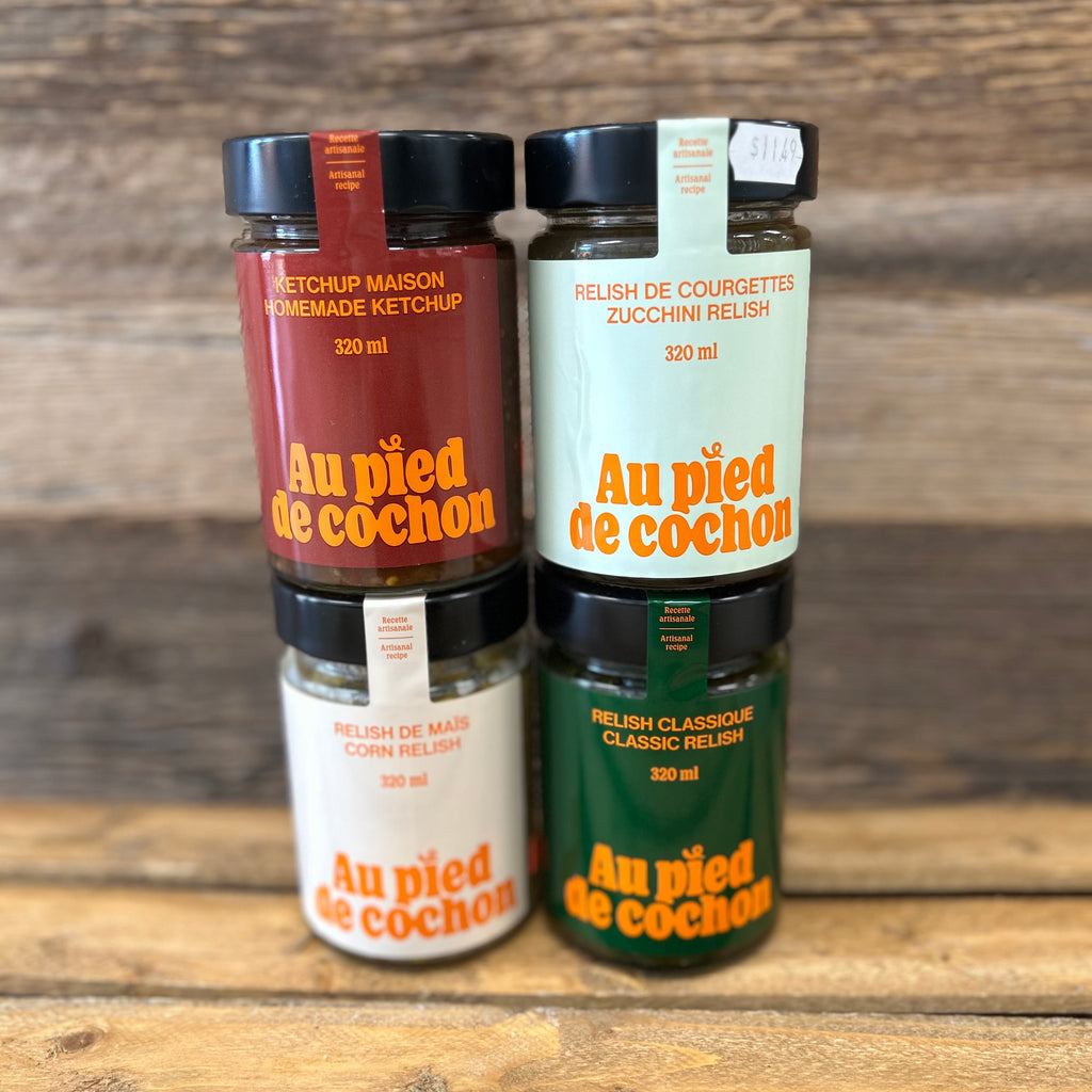 Four bottles of 'Au pied de cochon' condiments on a wooden surface with a wooden background.