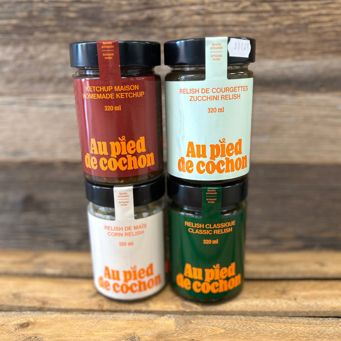 Four bottles of 'Au pied de cochon' condiments on a wooden surface with a wooden background.